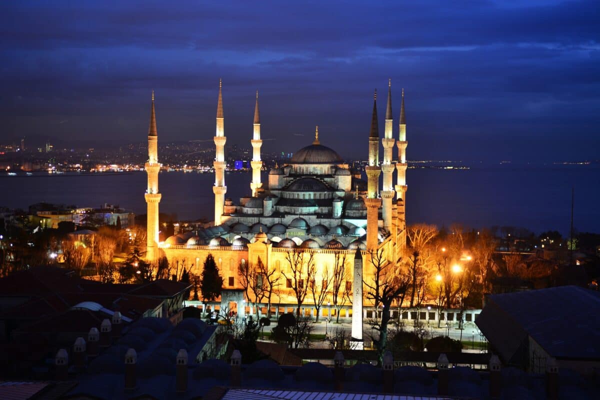 Foto da mesquita azul em Istambul na Turquia. Foto tirando durante a noite, ao fundo uma mesquita com várias torres e um domo ao meio. luzes em tons alaranjados
