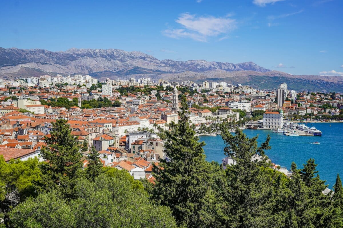 Vista de cima da cidade de Split na Croácia. Litoral cheio de casas banhado com mar azul e arvores a frente na foto.