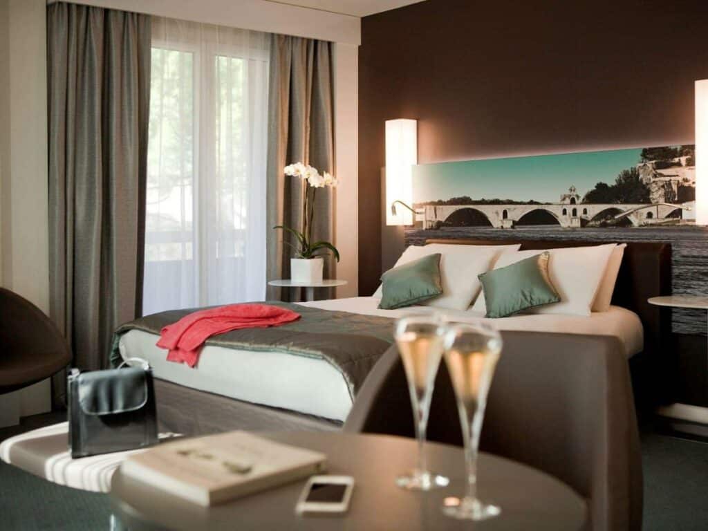 Quarto do Mercure Pont d’Avignon Centre, duas taças e champanhe e ao fundo a cama de casal branco com colcha marrom e almofadas verdes.