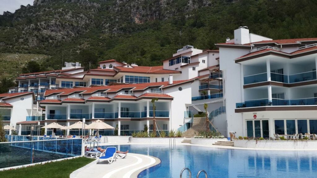 Foto externa do hotel Garcia Resort & Spa. Construção em branco e azul com telhados vermelhos e piscina a frente. Ao fundo montanha com pedra e mata verde