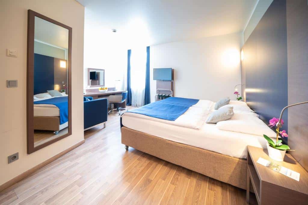 Quarto do Hotel Orangerie em Bruges na Bélgica. Quarto com cama de casal com roupa de cama branca e azul. Com grande espelho, poltrona e TV.