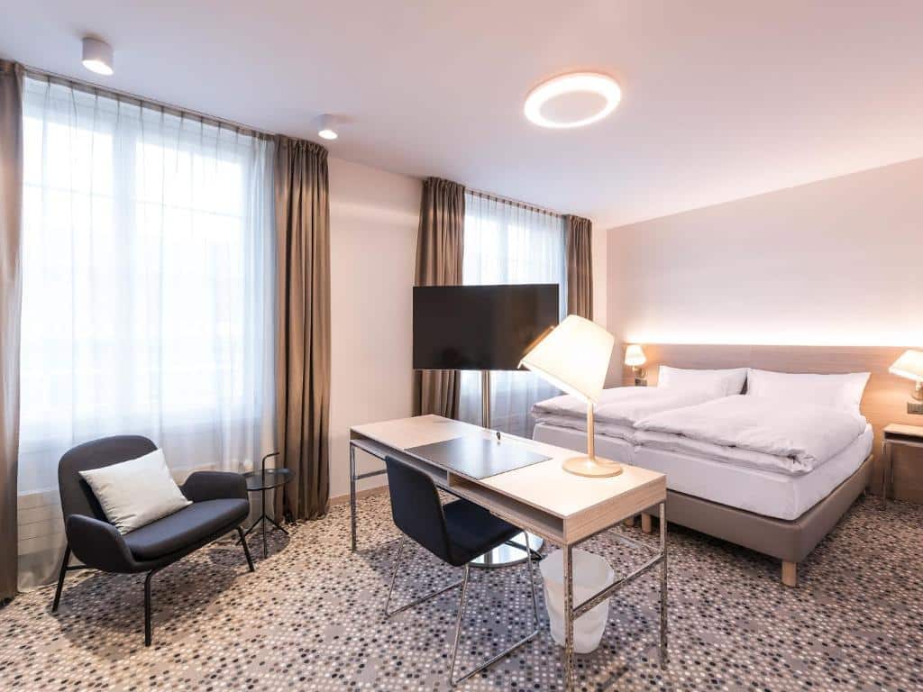 Quarto do hotel Hotel Savoy 
 em Berna na Suíça. Quarto em tons claros, branco e cinza. Cama de casal com roupa de cama branca. Duas janelas grandes com cortinas cinzas, mesinha de trabalho e poltrona.