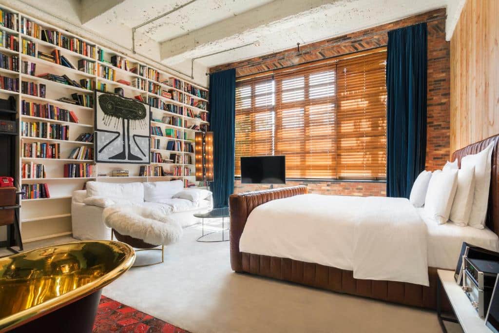 Quarto do Stamba Hotel, cama de casal com roupa de branca. Janela grande com cortina em madeira e em frente a cama vários livros.