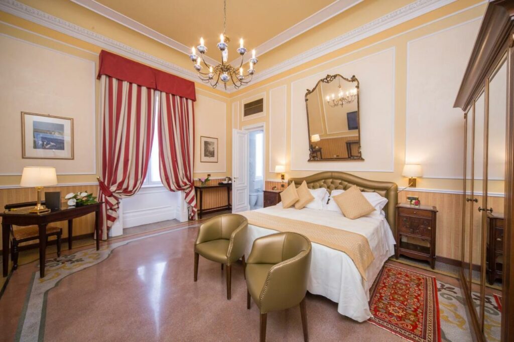 Quarto do hotel Bristol Palace em Genova - decoração em estilo clássico e com móveis de época, quarto em tons de branco, bege e vermelho com cama de casal, mesa de apoio e cortinas listradas de branco e vermelho.