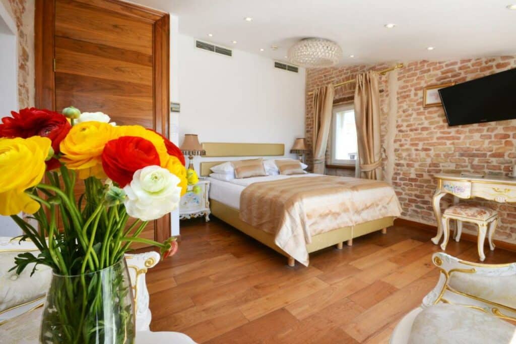 Quarto do Jupiter Luxury Hotel , cama de casal queen, com tons branco e bege. Tv e vaso com flores vermelhas e amarelas