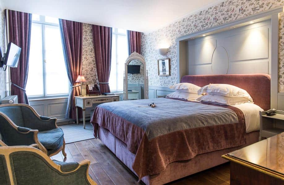 Quarto com cama de casal e almofadas, papel de parede em todo quarto, com cortinas proxima as janelas, espelho, poltronas, tv e luminarias representam um dos ambientes do hotel de orangerie em Bruges, na Belgica.