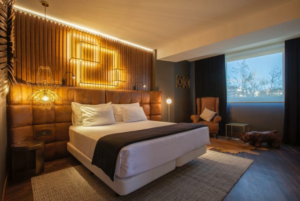 Quarto amplo com cama de casal, vista para o parque. Poltronas, criados e luminarias. Essa imagem representa um dos ambientes do Hotel Tres Reys em Pamplona