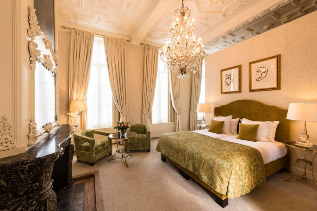 Quatro amplo com cama de casal, criados com luminarias, lustre de cristais, poltronas, lareira, cortinas classicas nas janelas. Essa imagem representa um dos ambientes do hotel Dukes' Palace Brugge em Flandres.