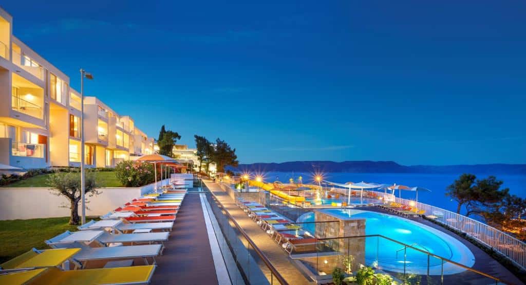 Entardecer no hotel Girandella Valemar Collection em Porec na Croacia. A imagem mostra as sacadas dos quartos que estão direcionadas para a área da piscina onde há espreguiçadeiras e luzes acesas. As sacadas também dão vista ao mar e montanhas ao fundo.