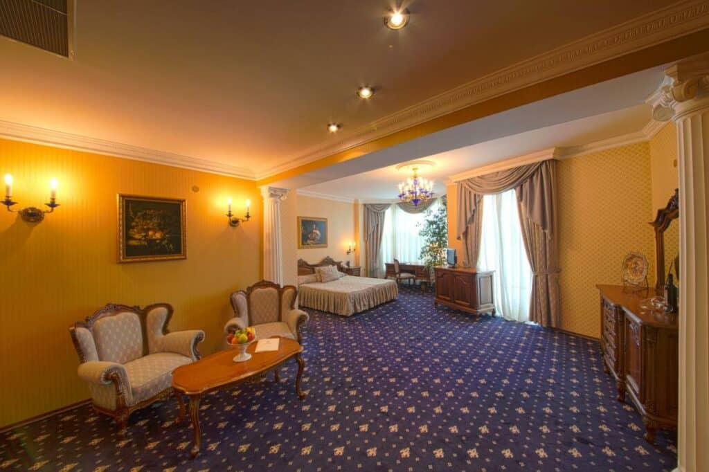 quarto espaçoso e sofisticado, com cama de casal, penteadeira, poltronas e mesa de centro. Janelas com cortinas classicas. Essa imagem representa um dos ambientes do hotel em Varna.