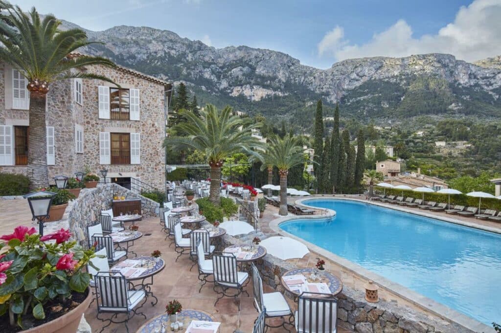 Belmond La Residencia em Mallorca - foto do hotel com paredes de pedra, piscina e cadeiras com almofadas listradas de azul e branco ao fundo montanhas e
