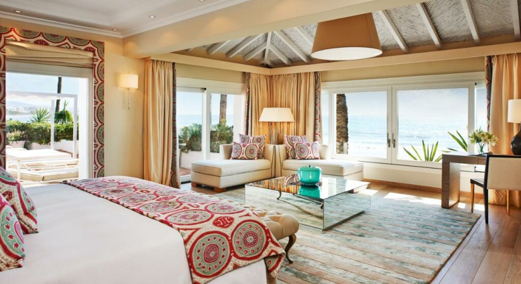 Quarto com cama de casal ampla e confortável, luminárias, sofas com almofadas. Bem iluminado, com varias janelas e vista privilegiada do mar. Essa imagem representa um dos hoteis de Marbella.