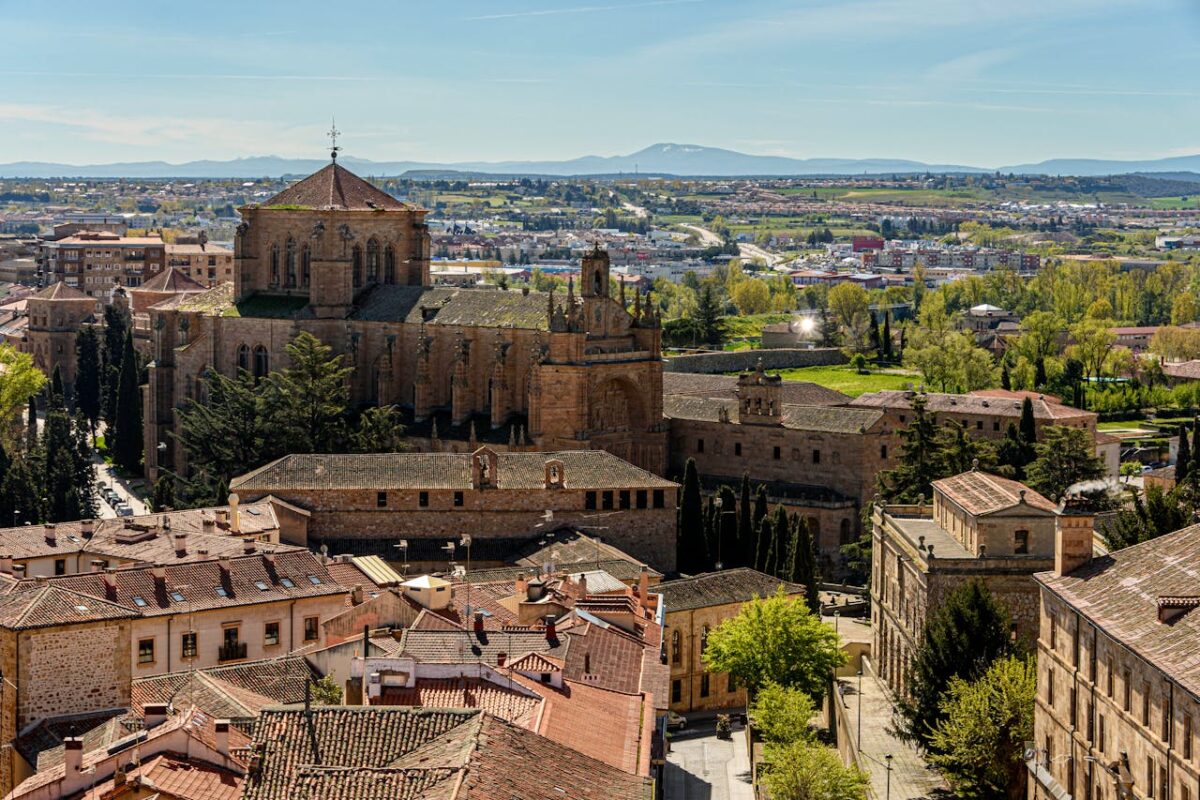 Vista aérea de uma antiga catedral de pedra cercada por árvores e edifícios, com uma paisagem urbana distante e montanhas ao fundo.