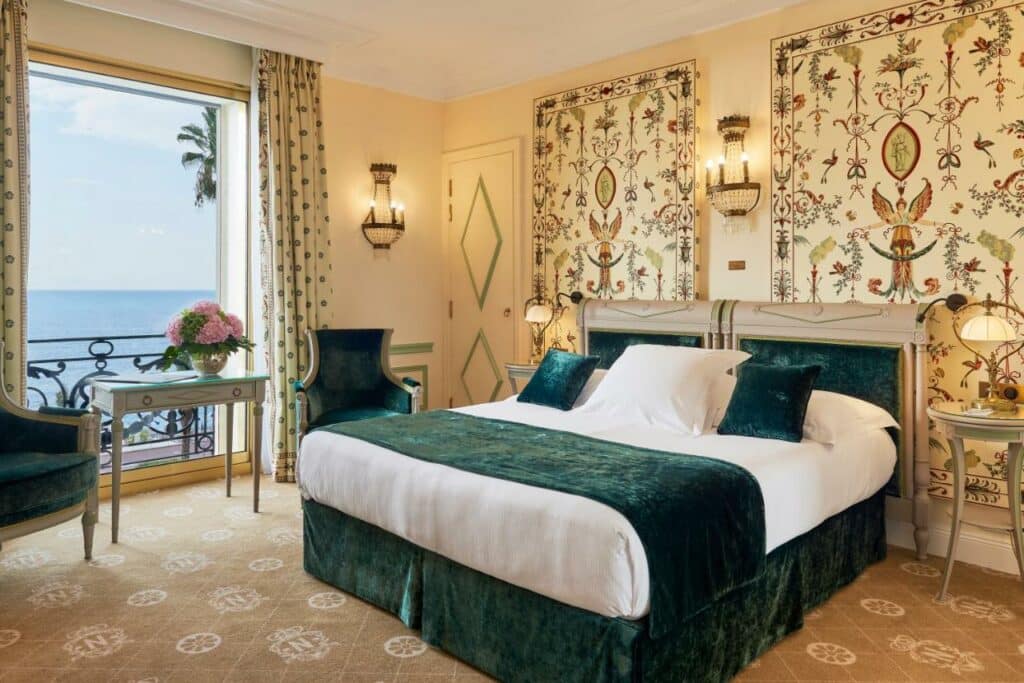 Quarto com cama de casal, bem decorado com papel de parede, luminarias, poltronas e uma linda vista para o mar. Essa imagem representa um dos ambientes do Hotel Le Negresco em Nice.