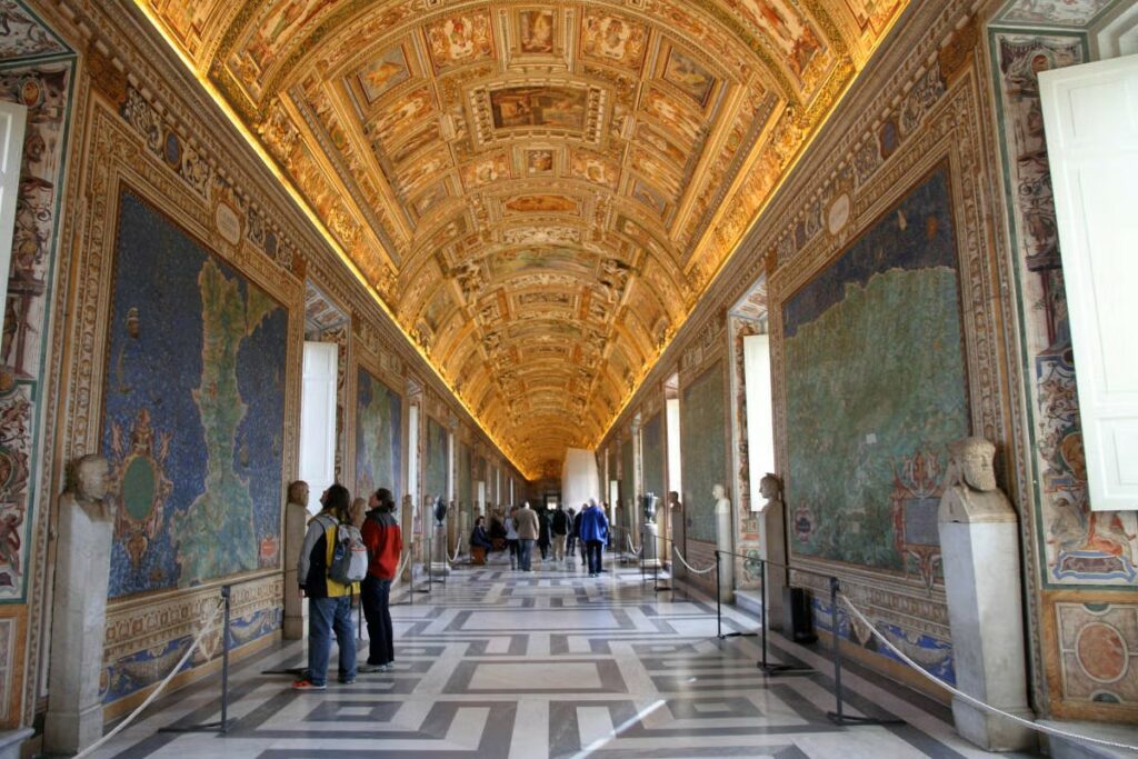 A imagem refere-se à Galeria dos Mapas, nos Museus do Vaticano. É possível observar o teto abobadado e decorado com afrescos deslumbrantes. Há mapas nas paredes que são do século XVI e representam as diferentes regiões da Itália. Há visitantes observando os mapas e as imagens do teto.