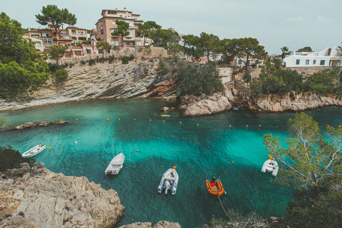 Praia de Peguera em Palma de Mallorca na espanha. Na imagem é possível observar alguns botes na água cristalina. e ao fundo um paredão rochoso com construções em cima.