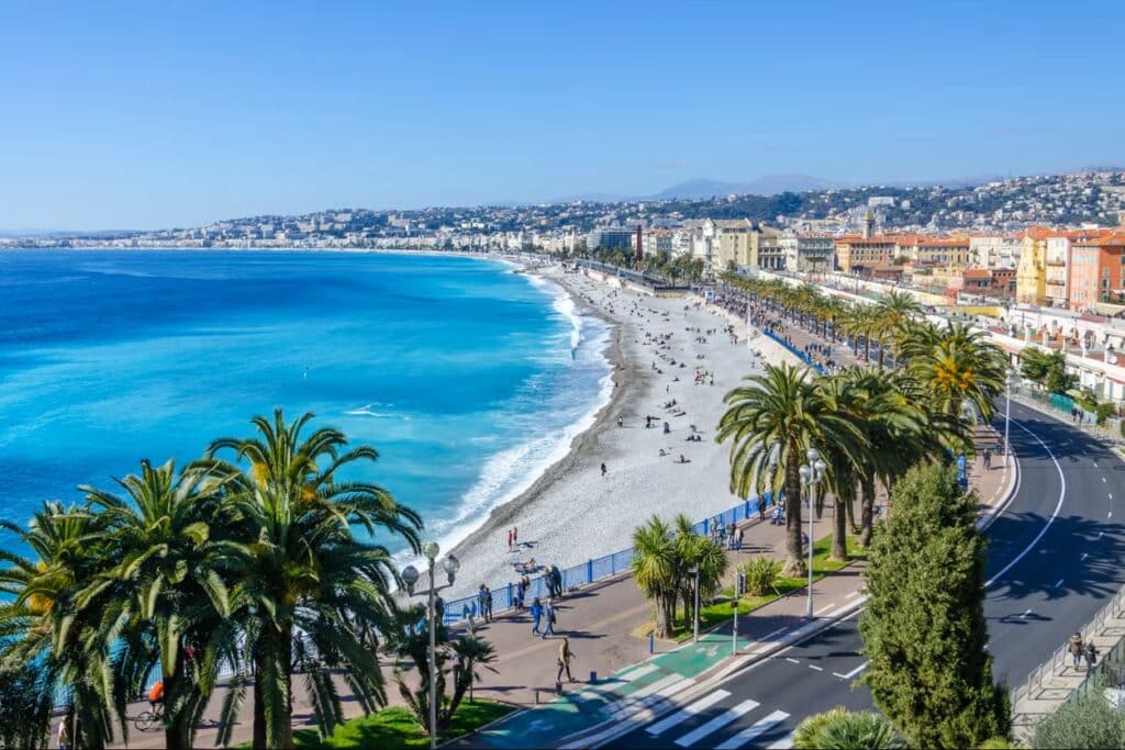 Vista panorâmica da orla marítima da cidade de Nice. É possível observar o mar, uma faixa de areia, coqueiros ao longo da orla e vista da cidade ao fundo.