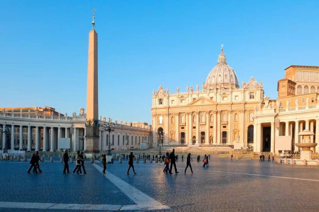 Praça de São Pedro na cidade do Vaticano, onde há pedestres caminhando com roupas de frio, durante o dia. No centro da fonte, ergue-se um imponente obelisco. Ele é alto e esguio. Ao fundo observa-se a Basílica de São Pedro.