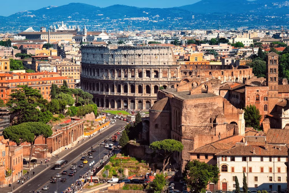Na imagem é possível observar o exterior do Coliseu na Cidade de Roma, e a cidade ao fundo e entorno o Coliseu.