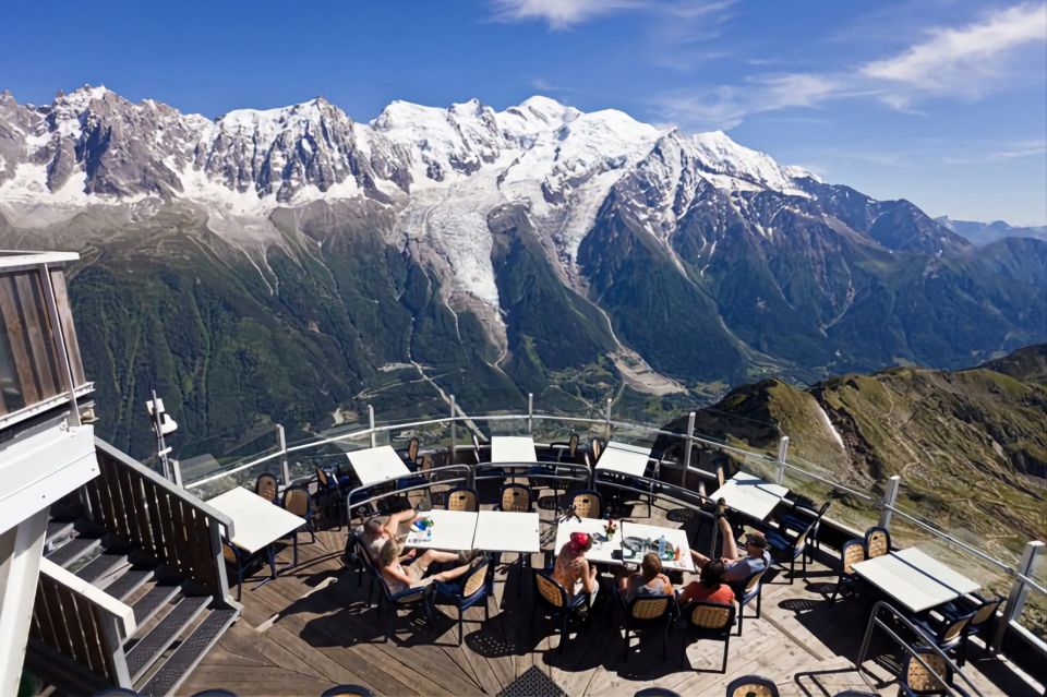 restaurante nos Alpes Franceses, na imagem há montanhas ao fundo com neve ao topo, e na parte inferior da imagem há mesas de um restaurante e algumas pessoas com roupas de inverno sentadas.