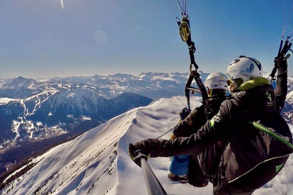 A imagem mostra duas pessoas saltando de parapente em Chamonix nos Alpes Franceses. Há montanhas ao fundo cobertas por neve.
