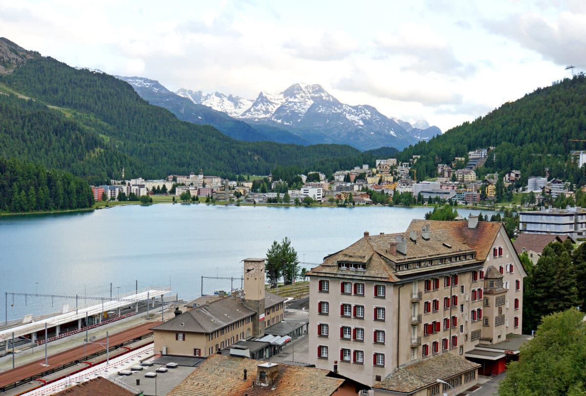 Lago cercado por montanhas e uma cidade com vários edifícios, incluindo uma estação de trem, sob um céu parcialmente nublado na cidade de St Moritz