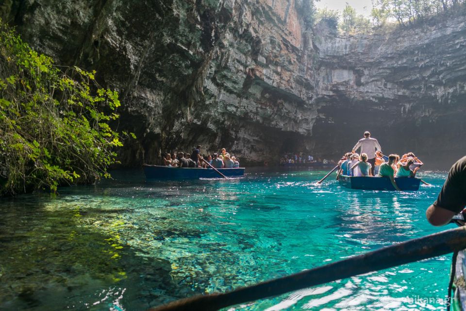 Excursão às cavernas da Kefalonia. Na imagem é possível observar uma gruta com entrada de luz natural e água cristalina. Há pequenos botes nas águas com visititantes.