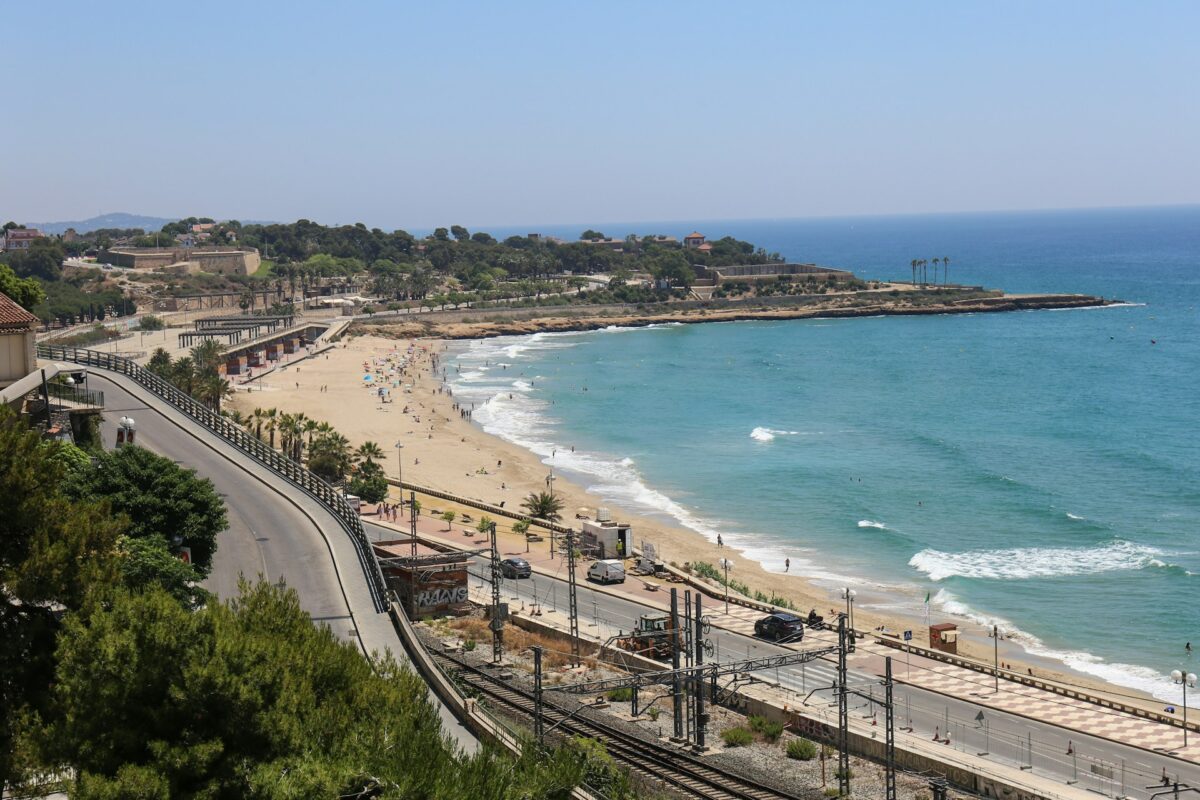 Paisagem costeira de Tarragona na Espanha, uma das cidades da Costa Dourada. Praia arenosa, oceano azul-turquesa e alguns visitantes espalhados. Uma estrada e árvores estão em primeiro plano.