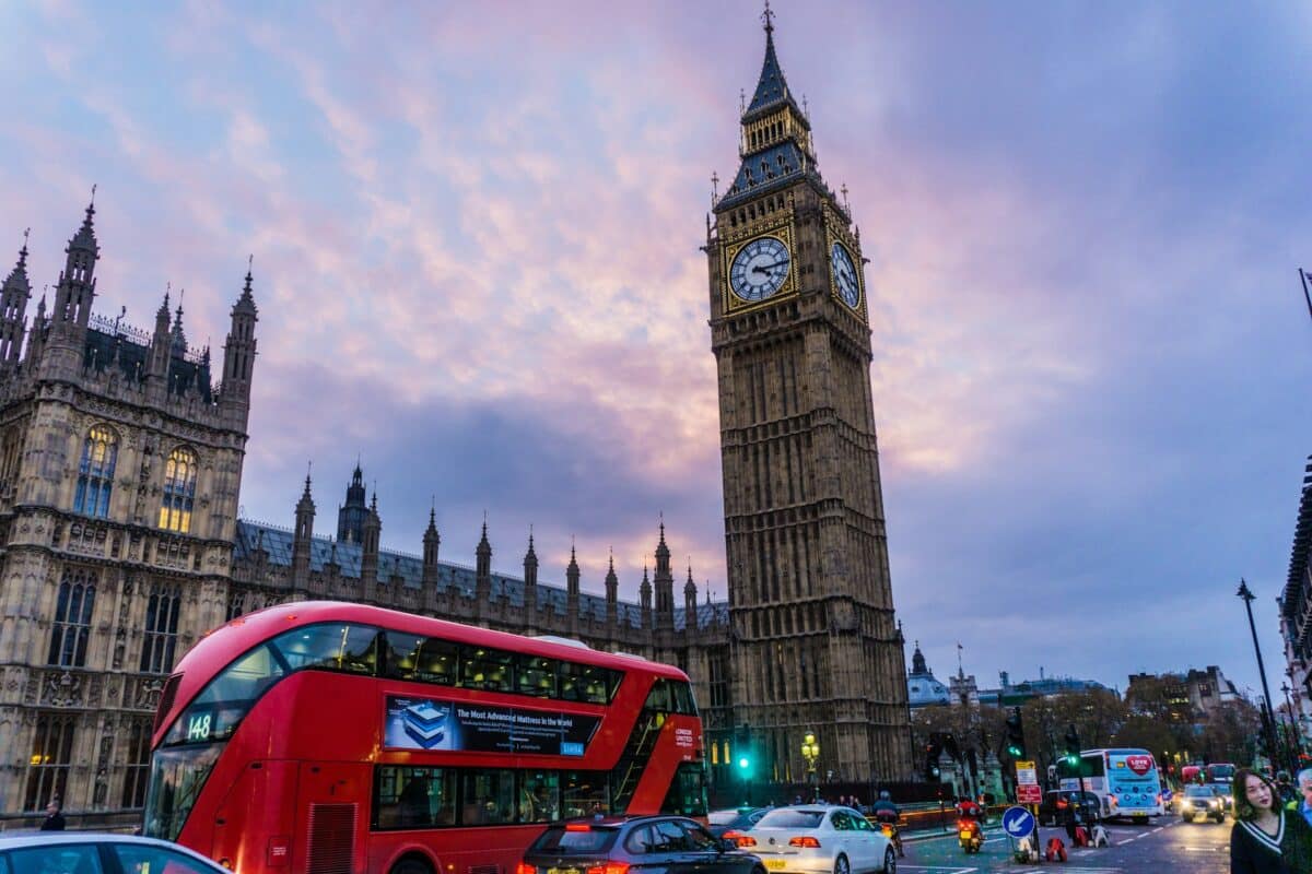 Um ônibus vermelho de dois andares e carros passam perto do Big Ben e das Casas do Parlamento em Londres ao anoitecer.