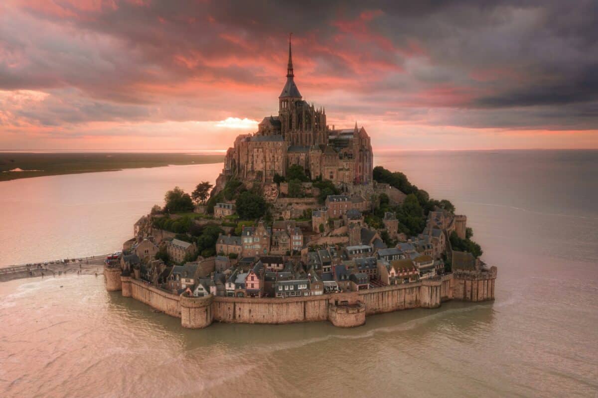 Vista aérea da ilha do Monte Saint-Michel com uma abadia ao pôr do sol sob nuvens dramáticas, cercada por água.