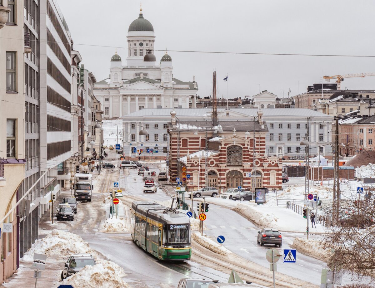 A imagem apresenta um trecho da cidade de Helsinque. A paisagem está toda coberta por neve, e há diversos prédios e monumentos antigos, nas ruas há carros e bondes elétricos, para representar aluguel de carros em Helsinque.