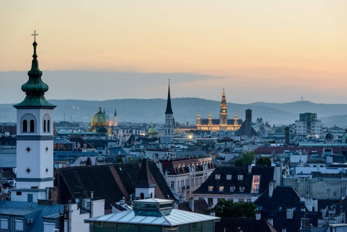 Paisagem urbana de Viena, na Áustria, ao anoitecer, com edifícios históricos, torres de igrejas e um céu suave ao pôr do sol ao fundo.