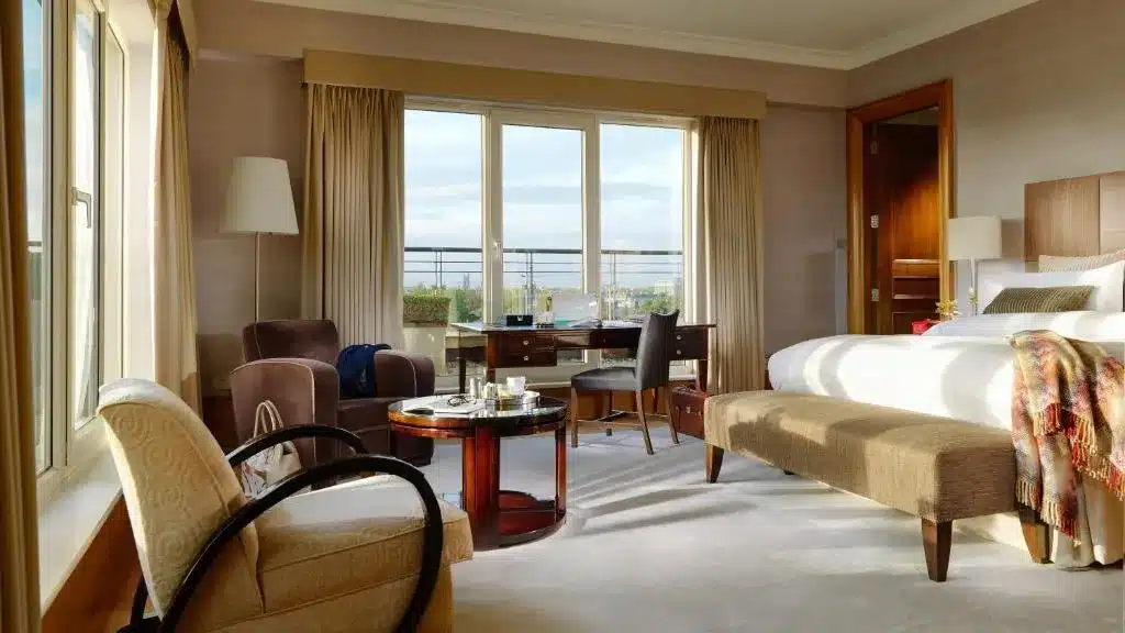 Quarto do Hebert Park Hotel and Park Residence, com uma cama grande, poltronas e uma mesa redonda. Uma escrivaninha de frente para uma janela com vista parcial para o exterior.