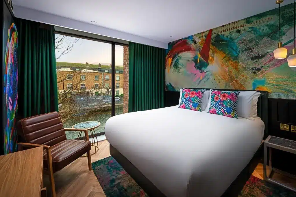 Um quarto moderno do NYX Hotel Dublin Portobello, com uma cama grande, arte abstrata vibrante na parede, uma cadeira de couro e uma janela com vista para um canal.