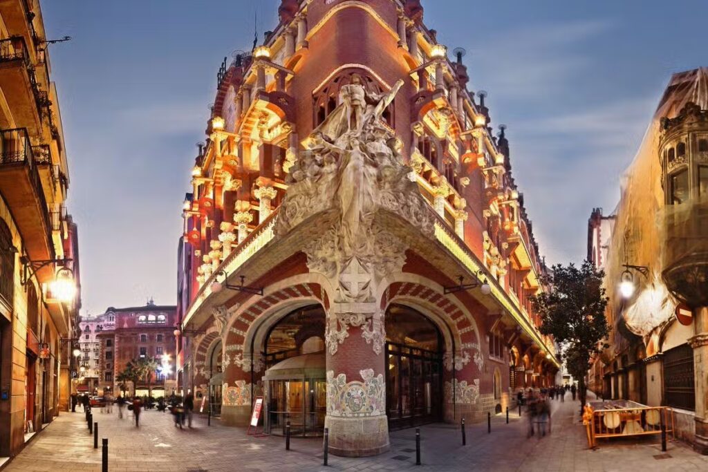 Palácio da música catalã, em Barcelona, Espanha. P´redio de esquina, muito ornamentado, com estátuas. Edifício rosa com detalhes em branco e luzes amarelas, com o céu azul de fim da tarde.