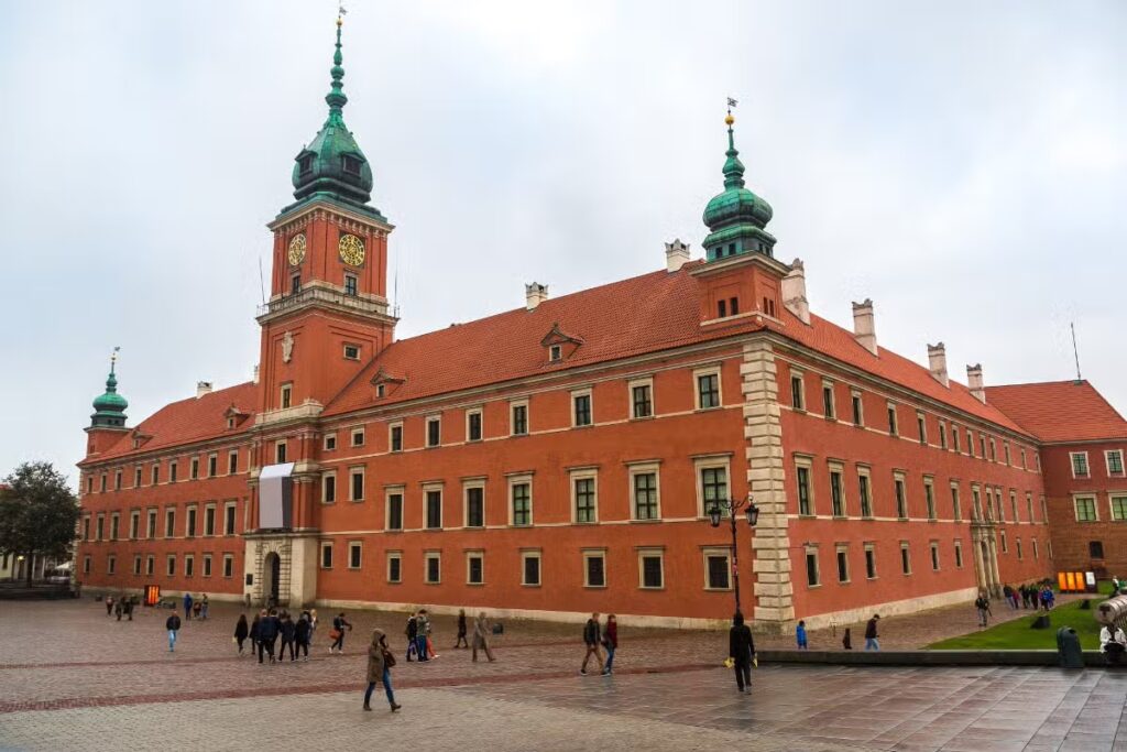 A imagem apresenta o Castelo Real de Varsóvia, em Varsóvia, na Polônia. Ele é um prédio retangular na cor laranja, com muitas janelas e algumas torres.