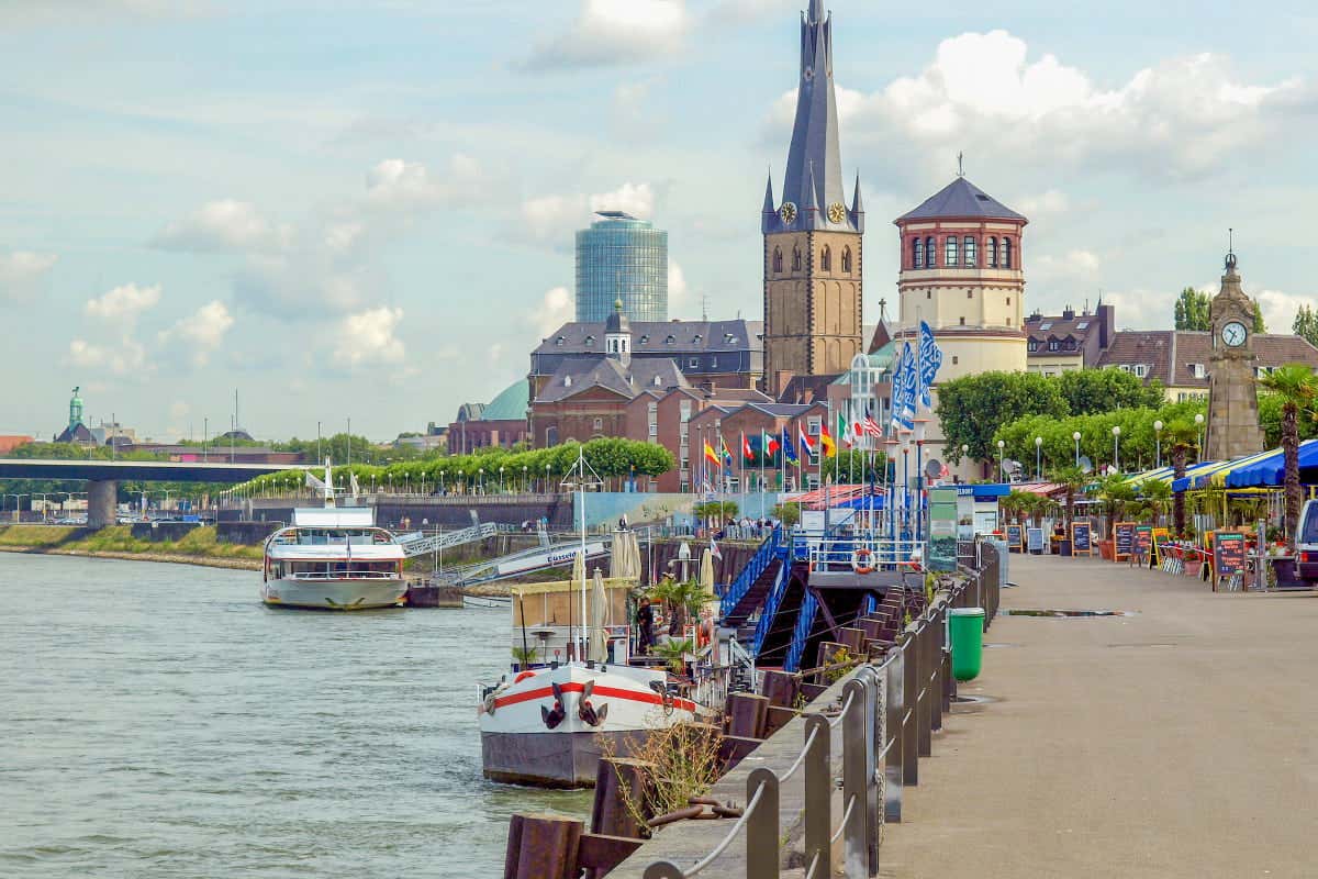 Cena de margem do rio Reno com barcos atracados ao longo do calçadão em Dusseldorf, Alemanha. Prédios históricos e uma torre de igreja são visíveis ao fundo.