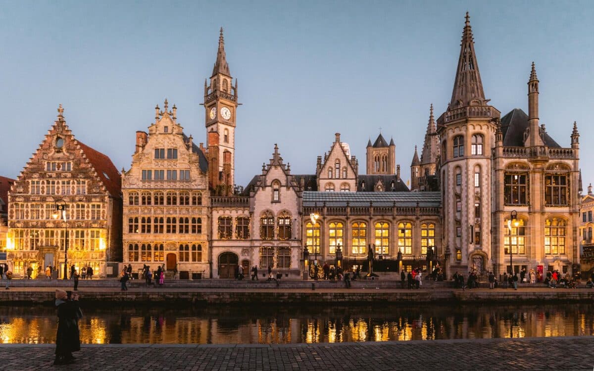 Uma fileira de edifícios históricos ornamentados em Gante, na Bélgica, com janelas iluminadas ao longo da margem de um rio durante o anoitecer, refletindo na água abaixo, para representar guia de viagem para a Bélgica