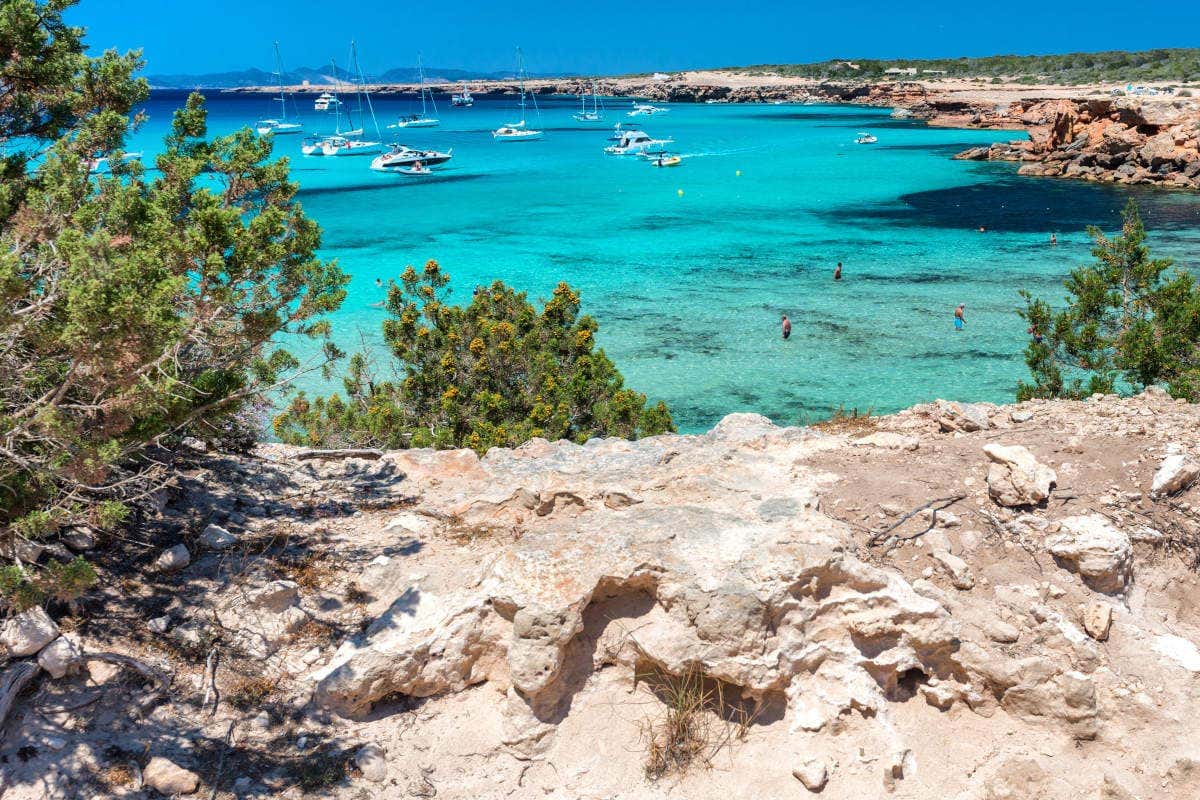 Costa rochosa com vista Cala Saona, em Formentera, Espanha, com água turquesa, vários barcos ancorados e nadadores em águas claras sob um céu azul. Vegetação visível.