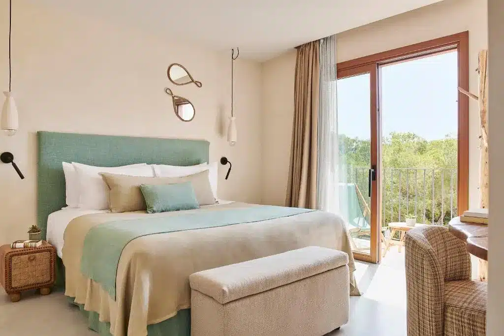 Quarto do hotel Teranka, em El Pujols, Formentera, Espanha, com uma cama grande, detalhes em bege e verde, um pequeno banco, cadeiras perto da janela e árvores verdes visíveis do lado de fora.