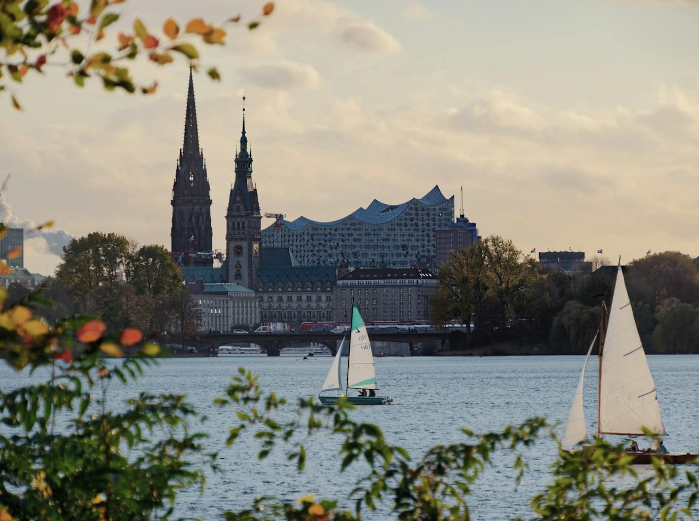 Veleiros no Rio Elbe, com uma paisagem urbana de Hamburgo, na Alemanha ao fundo, com duas torres e uma moderna sala de concertos sob um céu parcialmente nublado.