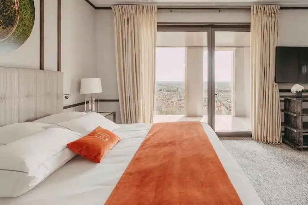 Quarto do hotel Villa Maïa, em Lyon, na França, com uma cama grande, corredor laranja e travesseiros brancos. Cortinas abertas para uma vista da sacada. Uma TV está montada na parede direita.