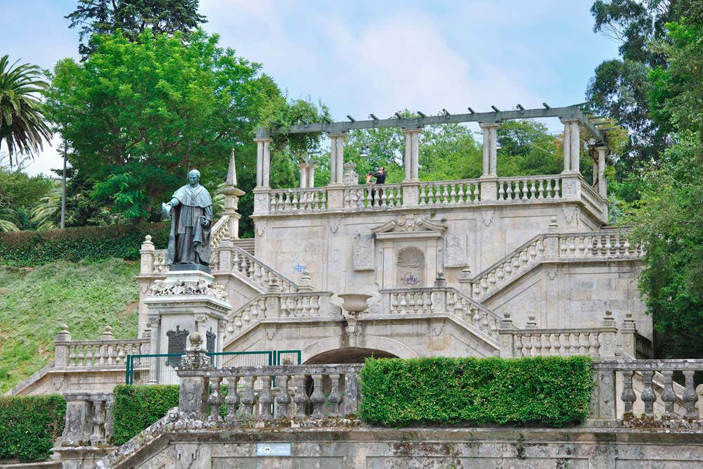Terraço de pedra com escada dupla e pérgola no topo, no Parque Alameda, em Santiago de Compostela, Espanha. Uma estátua fica à esquerda, cercada por vegetação e árvores.