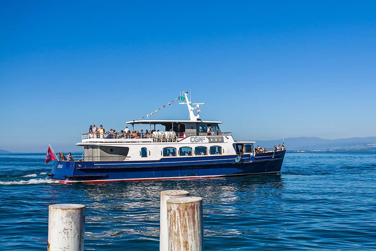 Barco azul e branco em um dia claro com pessoas a bordo, decorada com bandeiras coloridas, navegando em águas calmas, representando o passeio de barco pelo lago Léman.