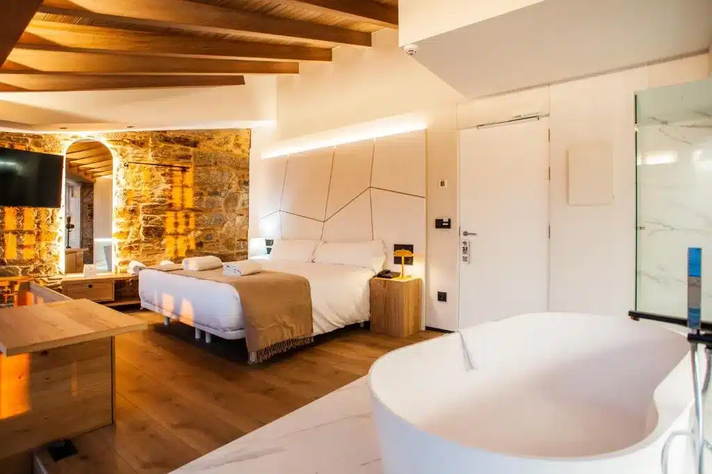 Quarto do Hotel Plaza Obradorio, em Santiago de Compostela, Espanha, com cama de casal, detalhes em madeira e pedra, TV e banheira independente. Iluminação suave cria um ambiente aconchegante.