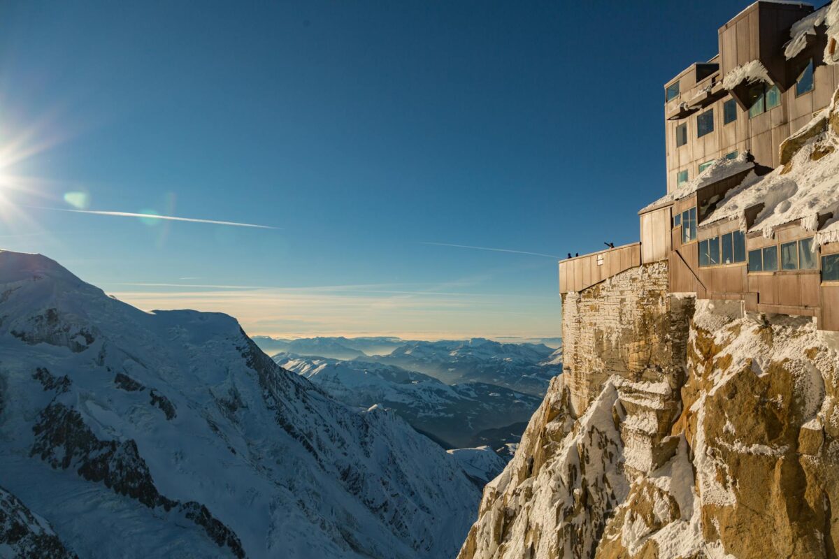 Paisagem de montanha nevada, em Chamonix, na França, iluminada pelo sol com um edifício no topo de um penhasco e um céu azul claro ao fundo.