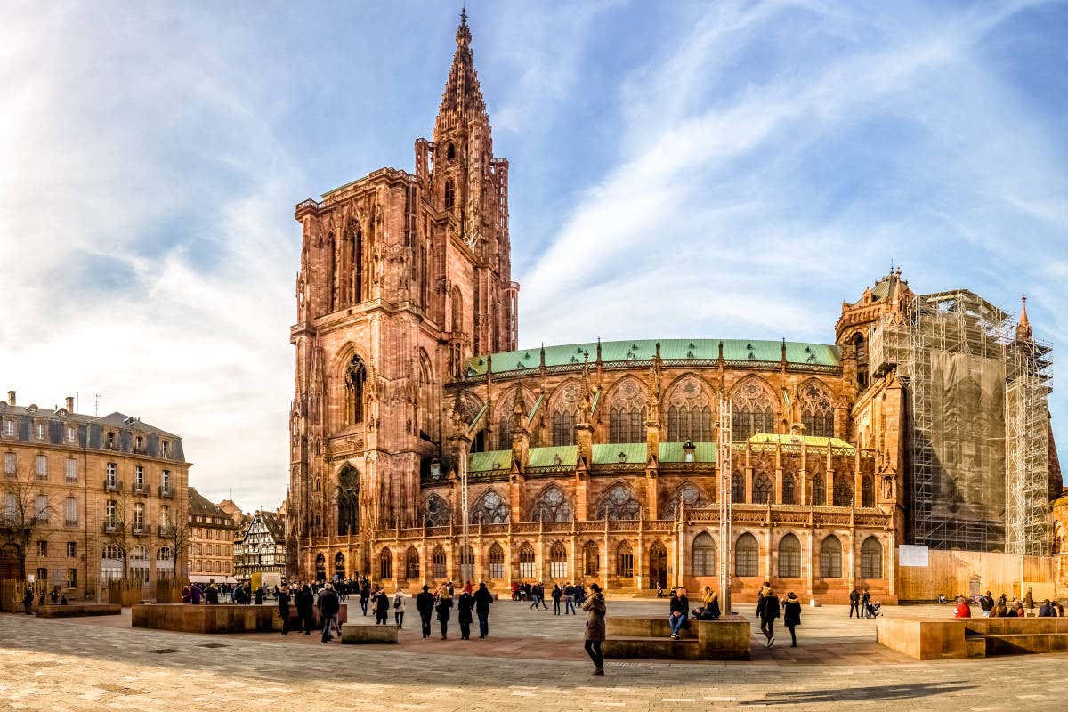 A Cadetral de Estrasburgo é uma grande catedral gótica com cantaria elaborada e uma torre alta, cercada por pessoas e edifícios adjacentes sob um céu azul.
