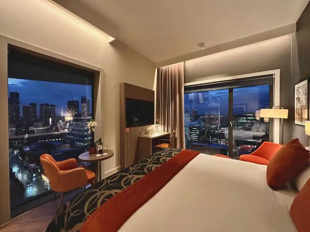 Quarto do Clayton Hotel Manchester City Centre, com uma cama, duas cadeiras laranjas, uma mesa, TV e grandes janelas mostrando uma paisagem urbana à noite, em Manchester, Inglaterra.