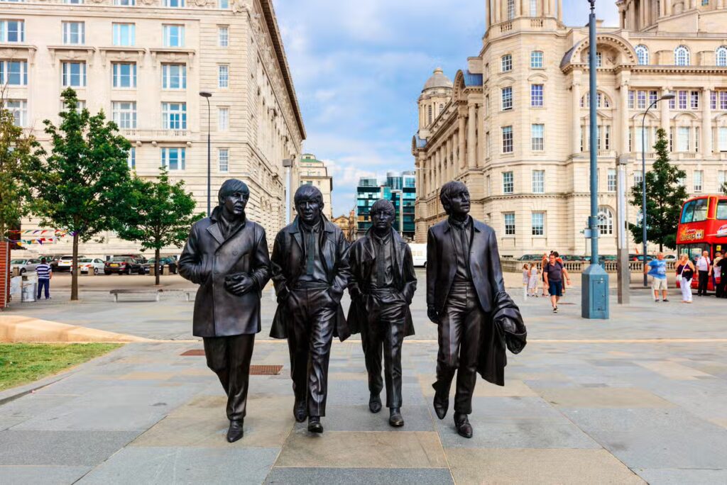 Estátuas de bronze de quatro homens da banda Beatles caminhando em frente a edifícios históricos em uma praça pública, com pessoas e árvores ao fundo, em Liverpool, na Inglaterra.