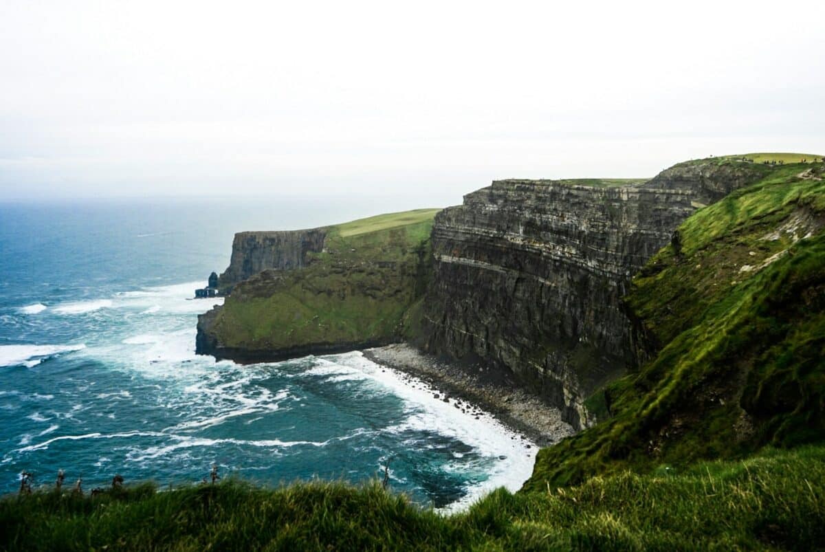 Cliffs of Moher com ondas do oceano sob um céu nublado, com grama verdejante no topo dos penhascos, formando uma paisagem costeira acidentada, na Irlanda.
